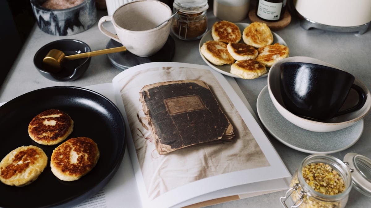 From above view of black plate with fresh cheese pancakes on opened book placed on table with various kitchenware in modern apartment